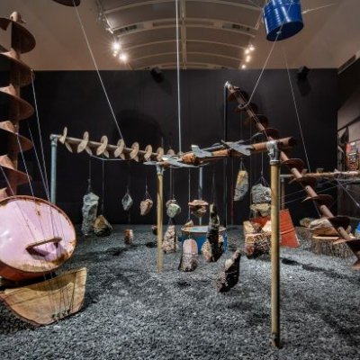 A room with dark walls is filled with an art installation of industrial mining equipment repurposed into musical instruments.